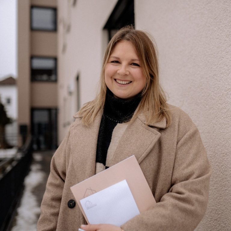 Lisa Lamparter, lächelnd vor einem Haus mit Mappe und Block in der Hand