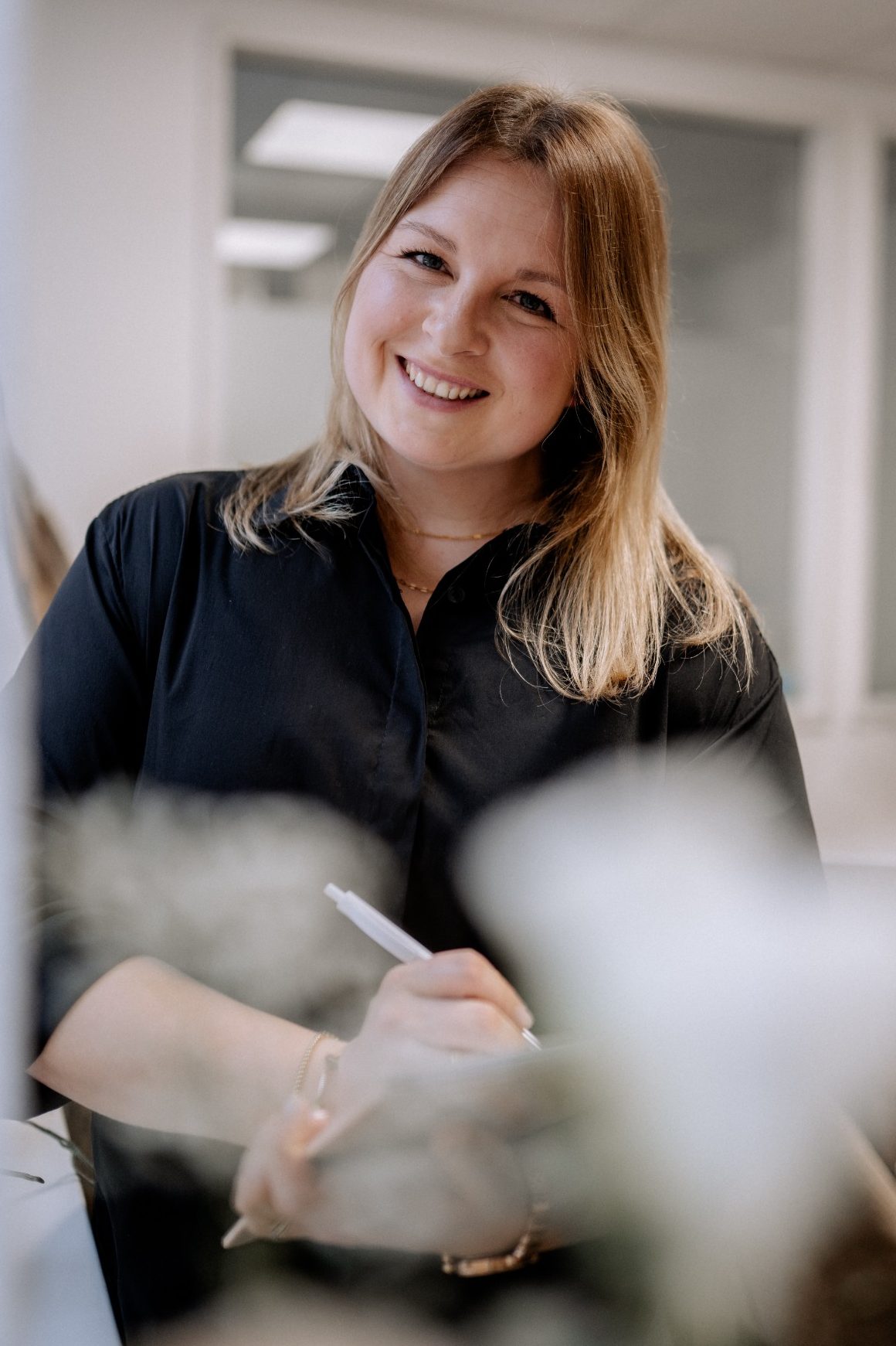 Lisa Lamparter mit schwarzer Bluse und Kugelschreiber in der Hand lehnt an der Wand und lächelt.